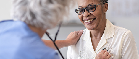 A woman wearing glasses smiles while a medical professional listens to her heartbeat with a stethoscope.