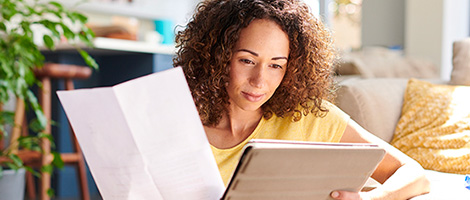 A woman is sitting on the floor at home while holding a piece of paper and looking at a device screen.